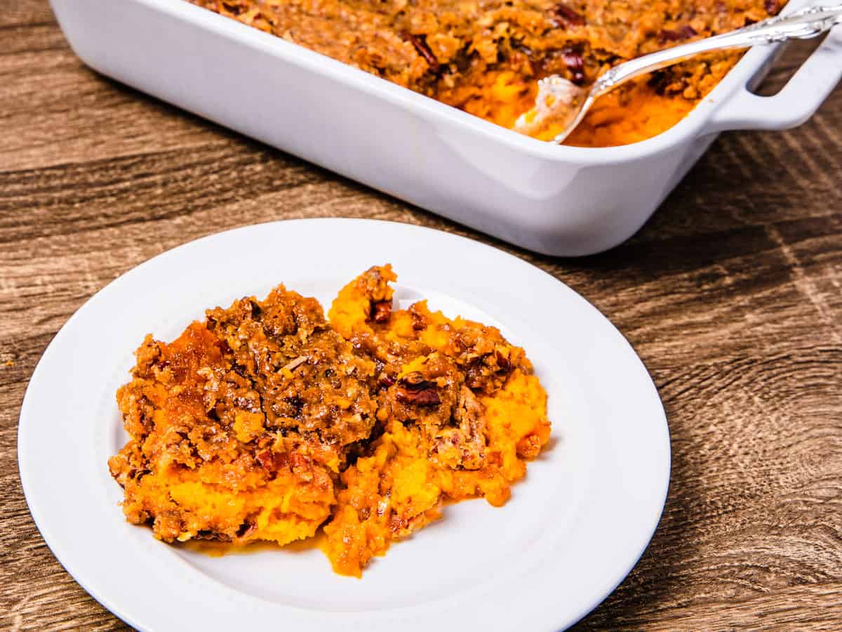 A serving of sweet potato casserole shown on a plate set on a table next to the baking dish. 