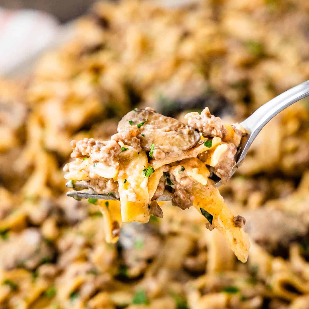 Close up of a fork with a bite of beef stroganoff held in front of a platter of the dish blurred in the background.