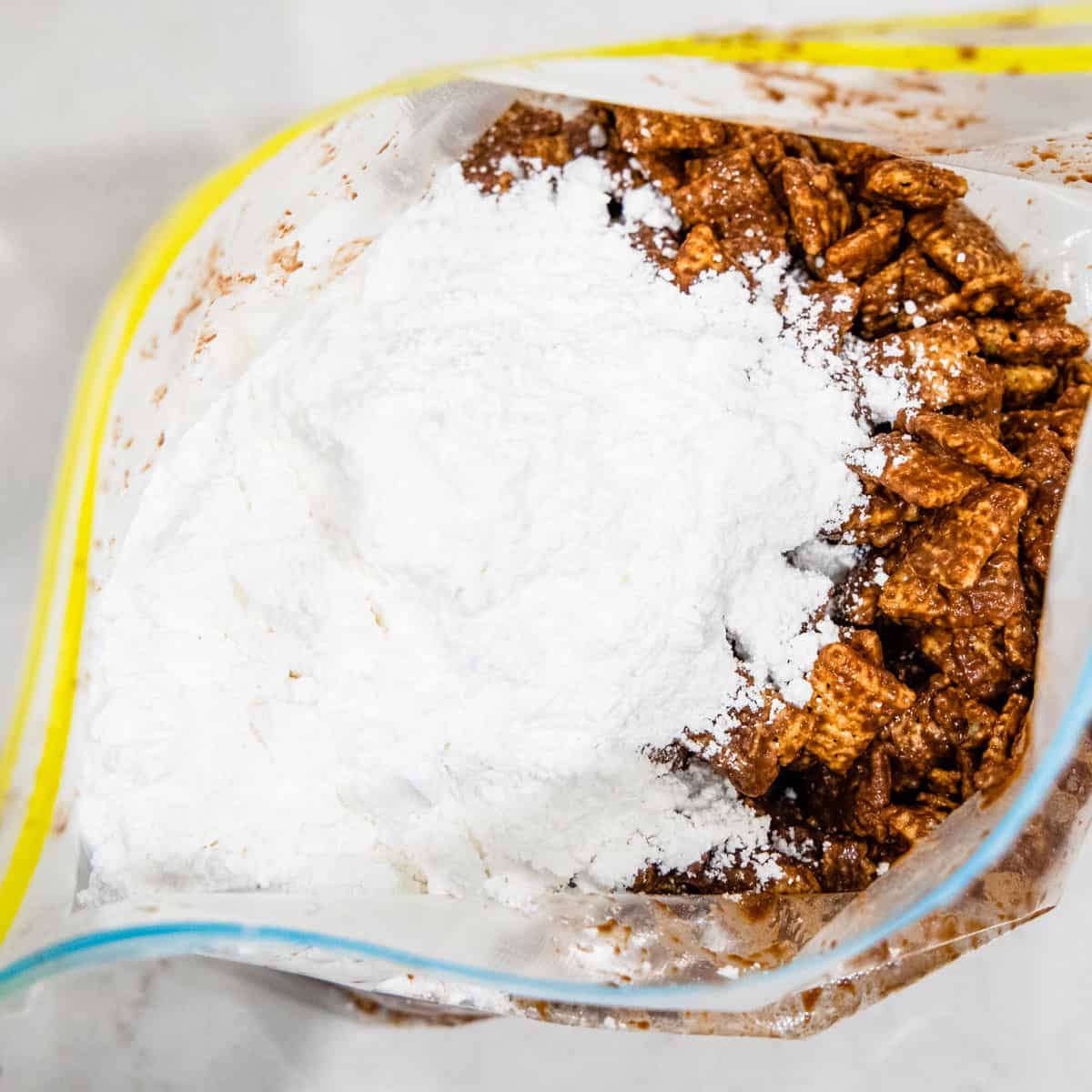 Chocolate-coated Chex cereal is shown in a large plastic bag with powdered sugar added in, before being mixed.