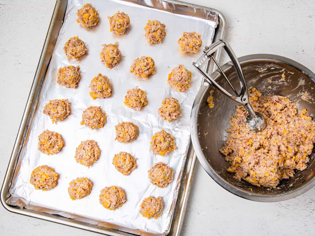 Sausage balls scooped onto a baking sheet sitting next to a bowl of the remaining sausage mixture.