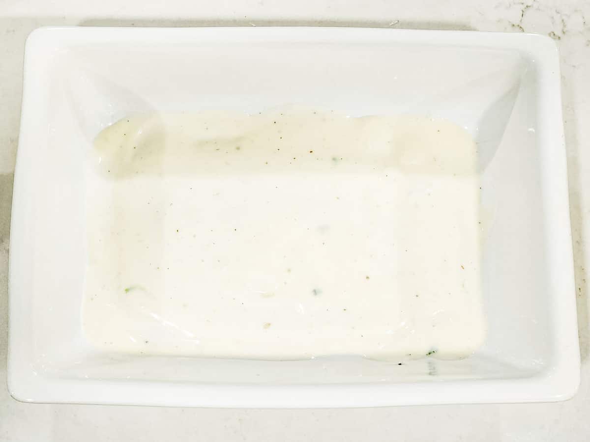 A baking dish shown with a cup of white sauce spread on the bottom.
