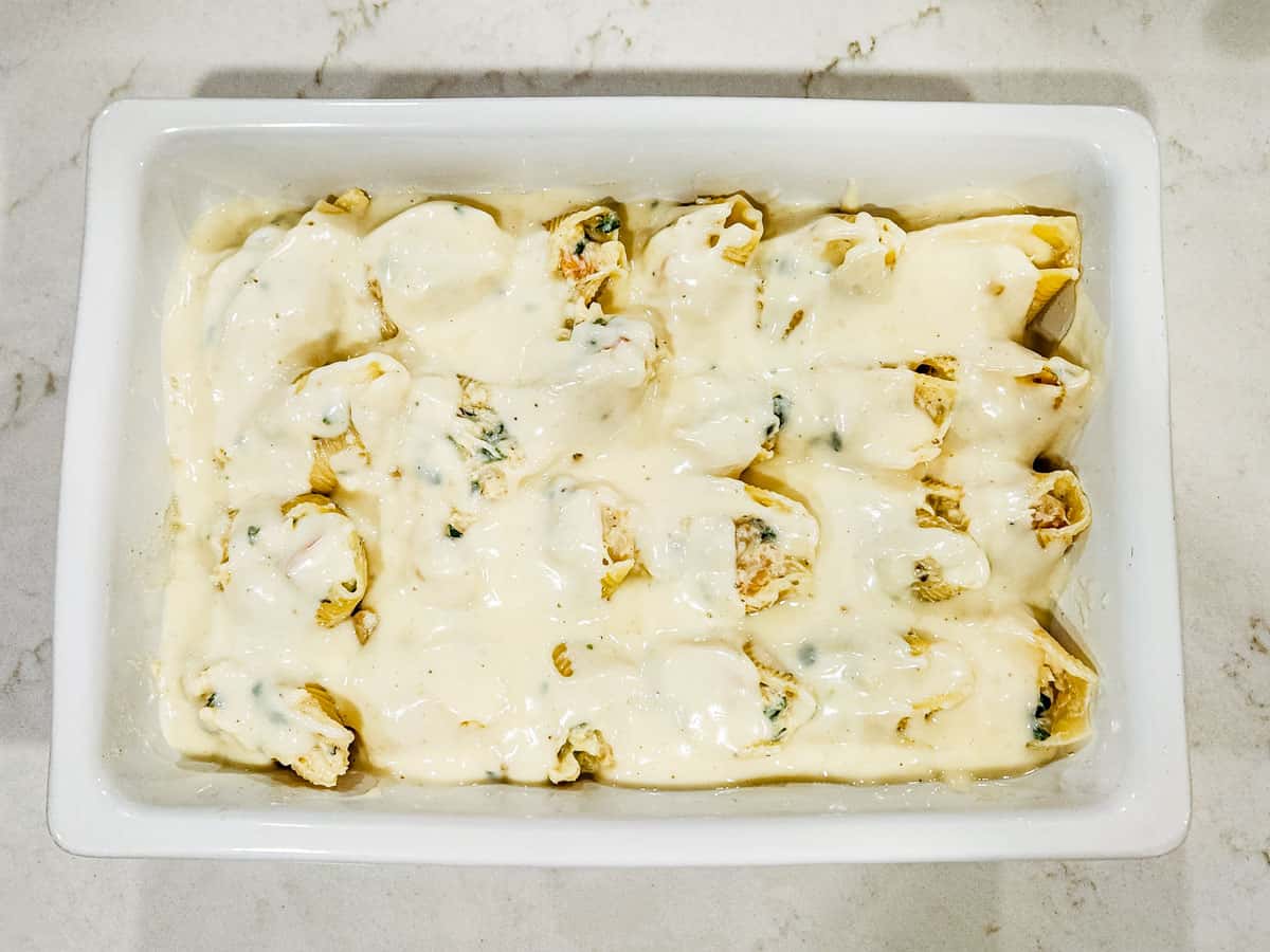 Stuffed pasta shells in a baking dish topped with white sauce. 