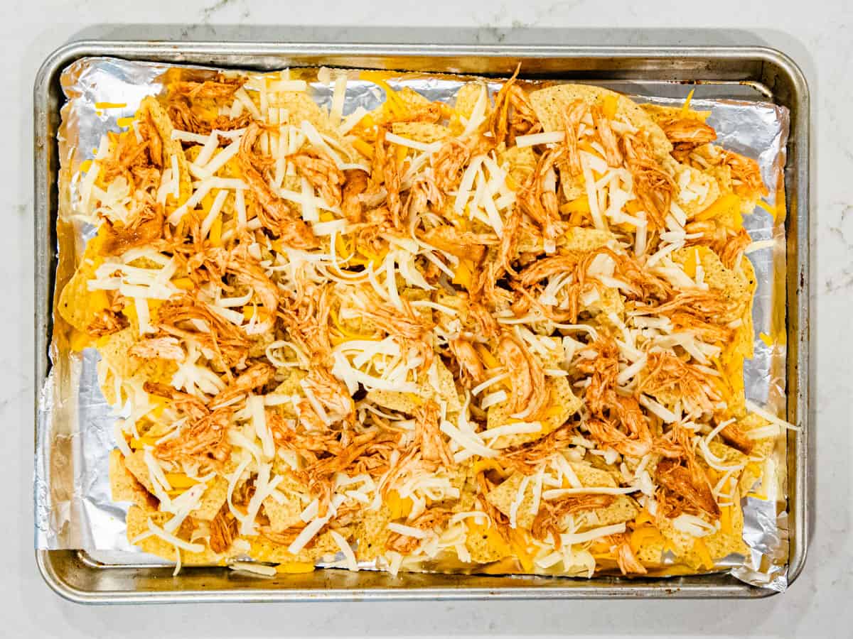 Tortilla chips topped with shredded cheese and BBQ chicken on a sheet pan before being baked. 