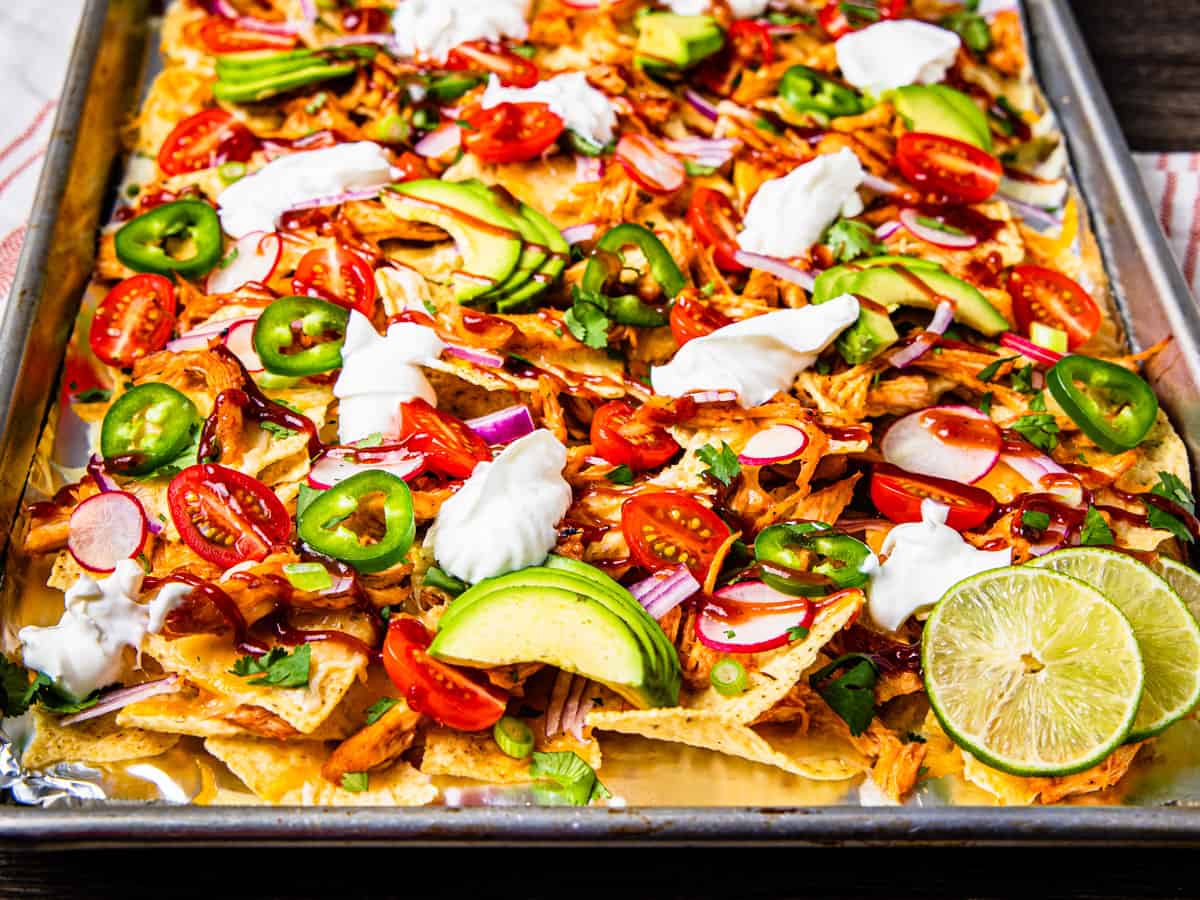 A sheet pan of barbecue chicken nachos topped with sliced avocado, jalapeños, cilantro, tomatoes, and sour cream. 