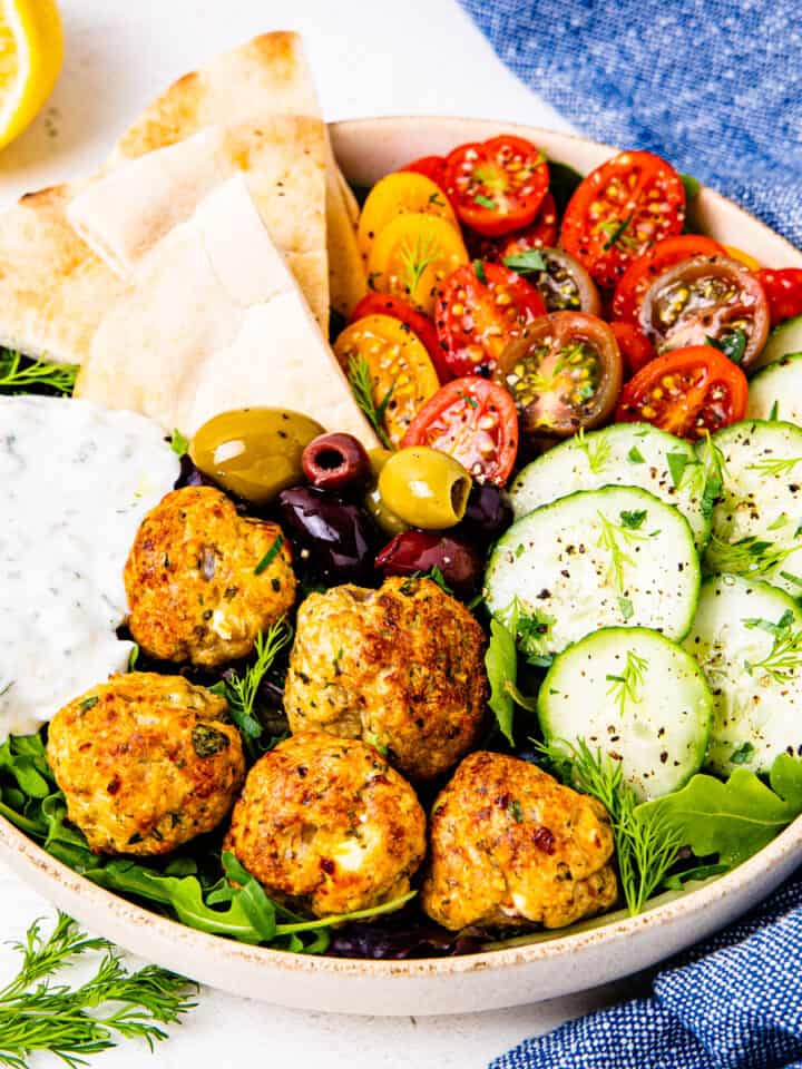 A bowl of Greek salad topped with chicken meatballs and tzatziki sauce.