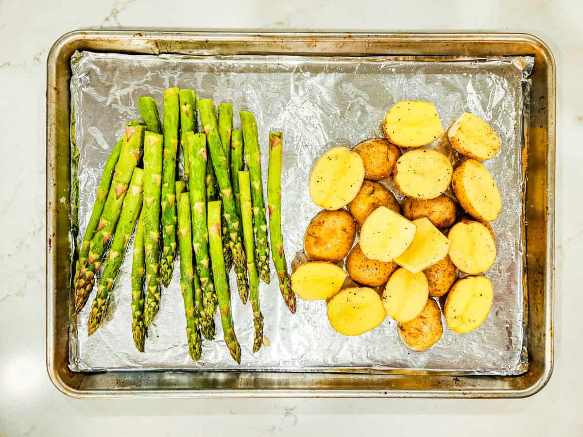 A pile of halved potatoes and a stack of asparagus shown on a sheet pan. 