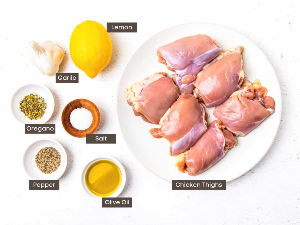 A plate of boneless skinless chicken thighs set beside individual ingredient bowls of salt, pepper, oregano, olive oil, a lemon, and a head of garlic.