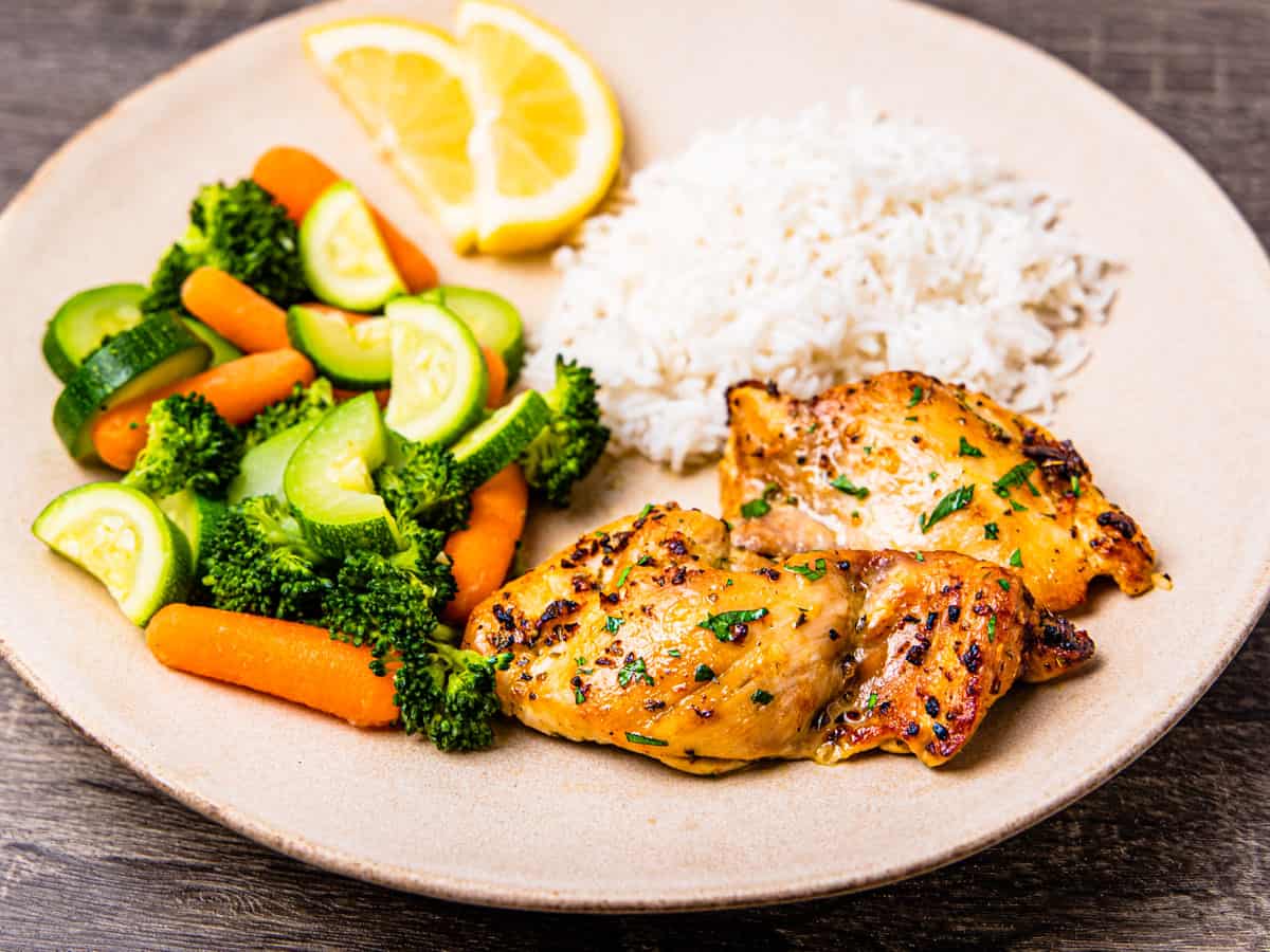 A plate with steamed rice, mixed vegetables, and boneless chicken thighs.