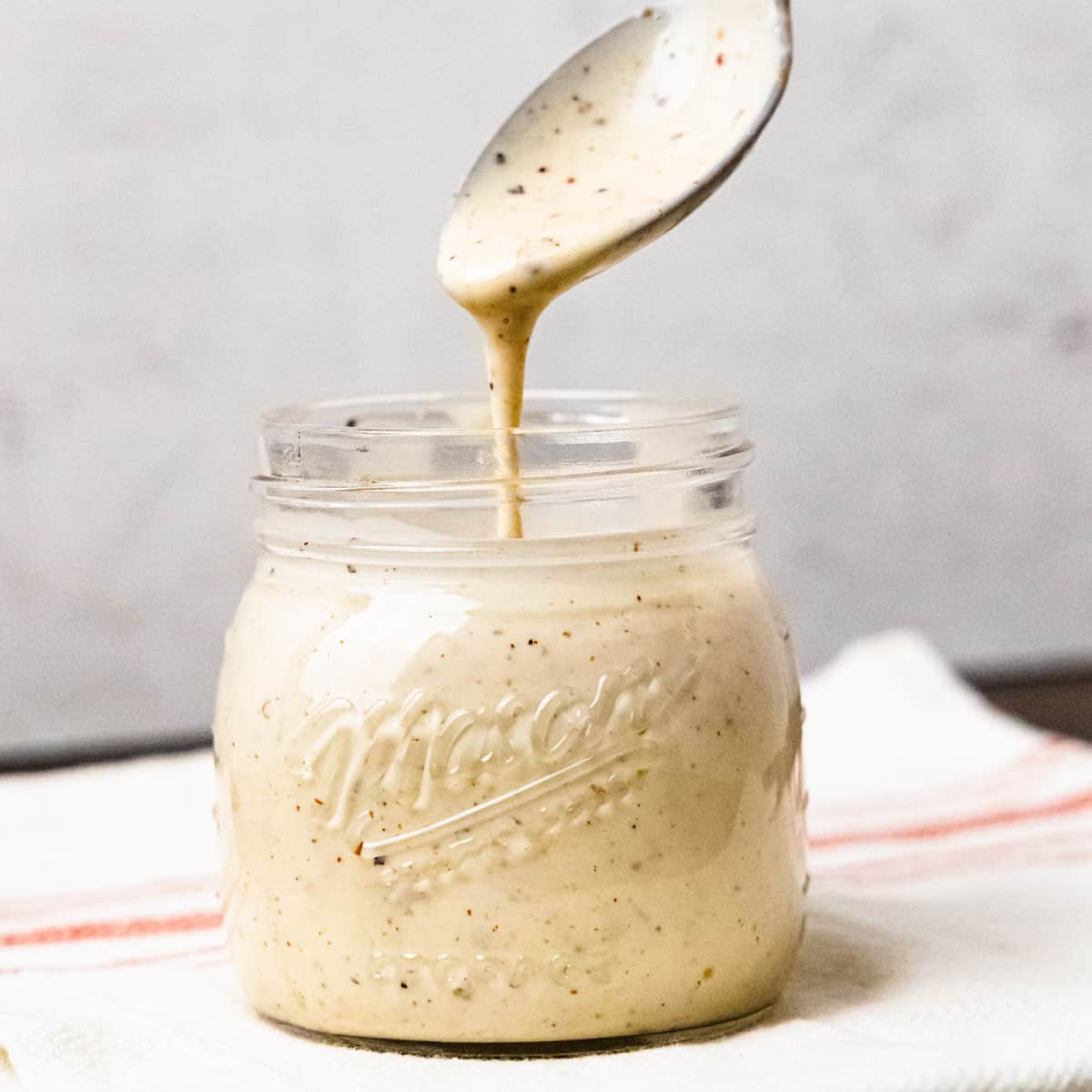 A spoon held above a jar of Alabama white barbecue sauce with sauce drizzling off the spoon into the jar.