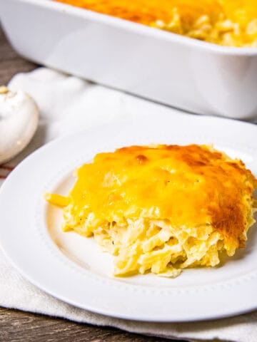 A serving of cheesy hashbrown casserole on a small plate sitting on a table next to a casserole dish.
