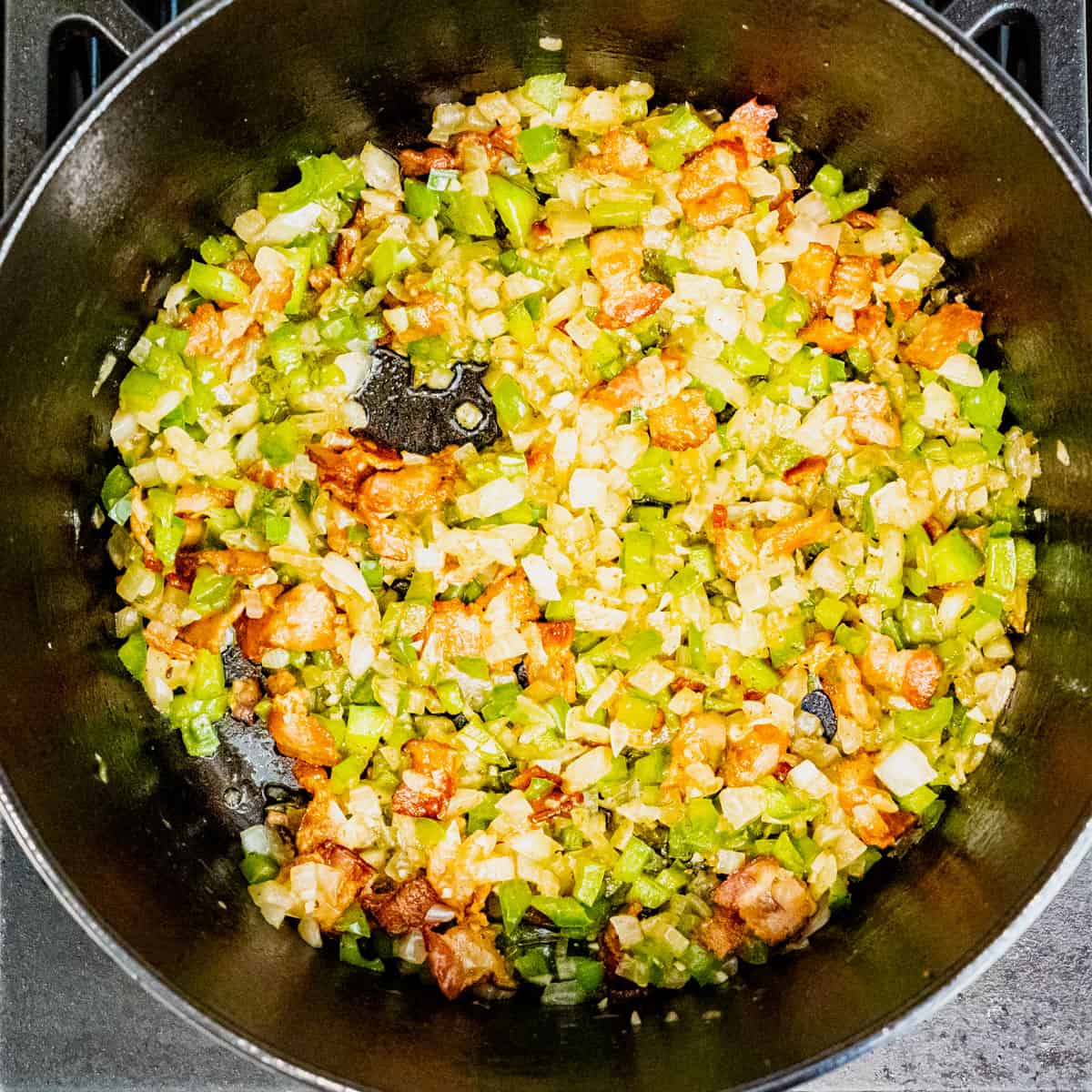 Sautéed bacon, onion, and green pepper cooking on a stovetop in a cast iron pot.