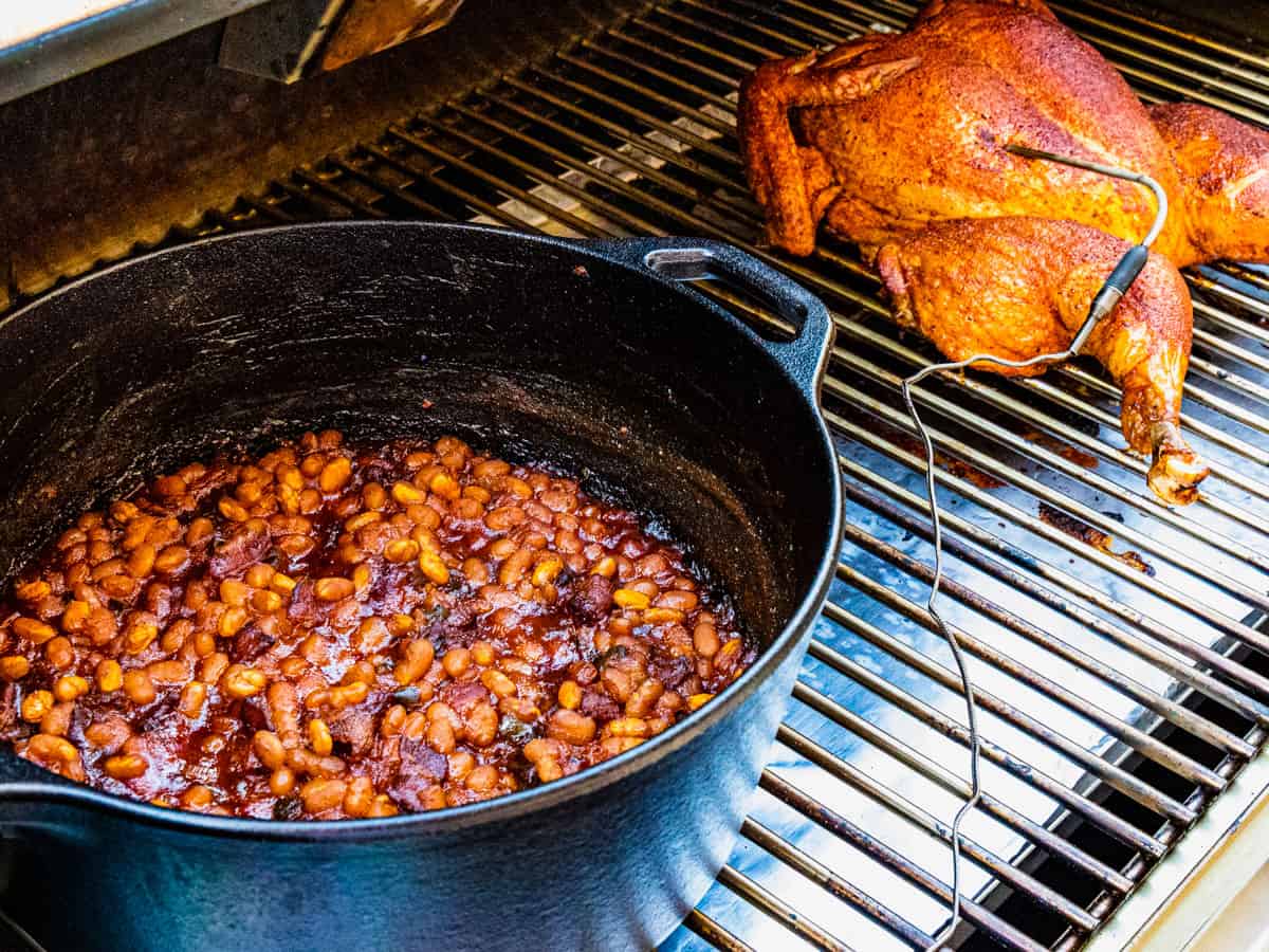 A large pot of baked beans and a whole chicken cooking on a smoker.