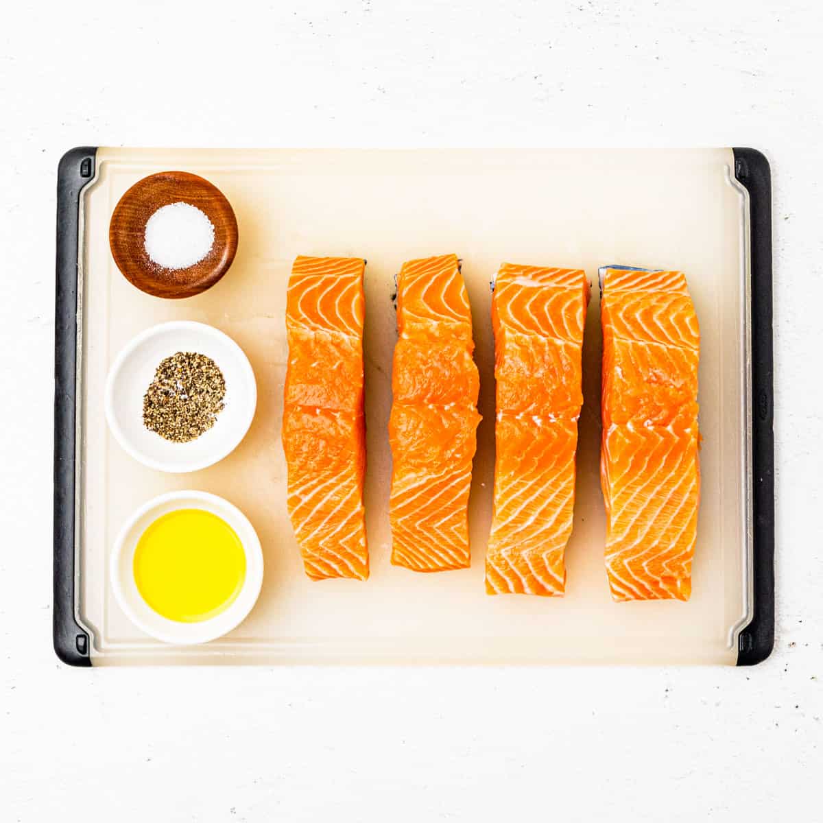 Salmon fillets on a cutting board next to small pinch bowls of olive oil, salt, and pepper.