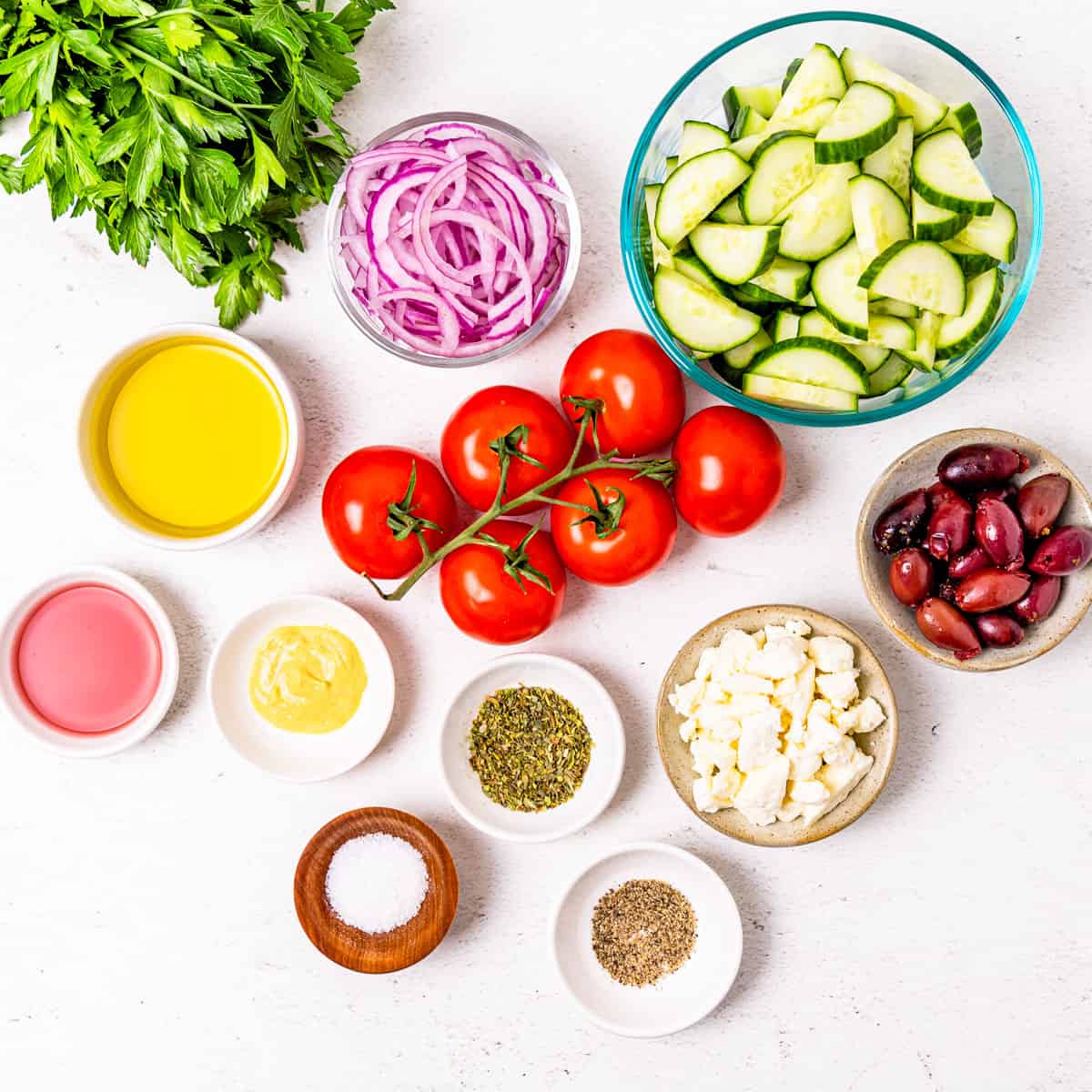 Ingredients for traditional Greek salad and a Greek vinaigrette dressing arranged in individual bowls on a countertop.