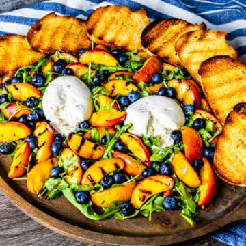 Grilled peaches with burrata, arugula, and blueberries served on a platter with grilled bread.