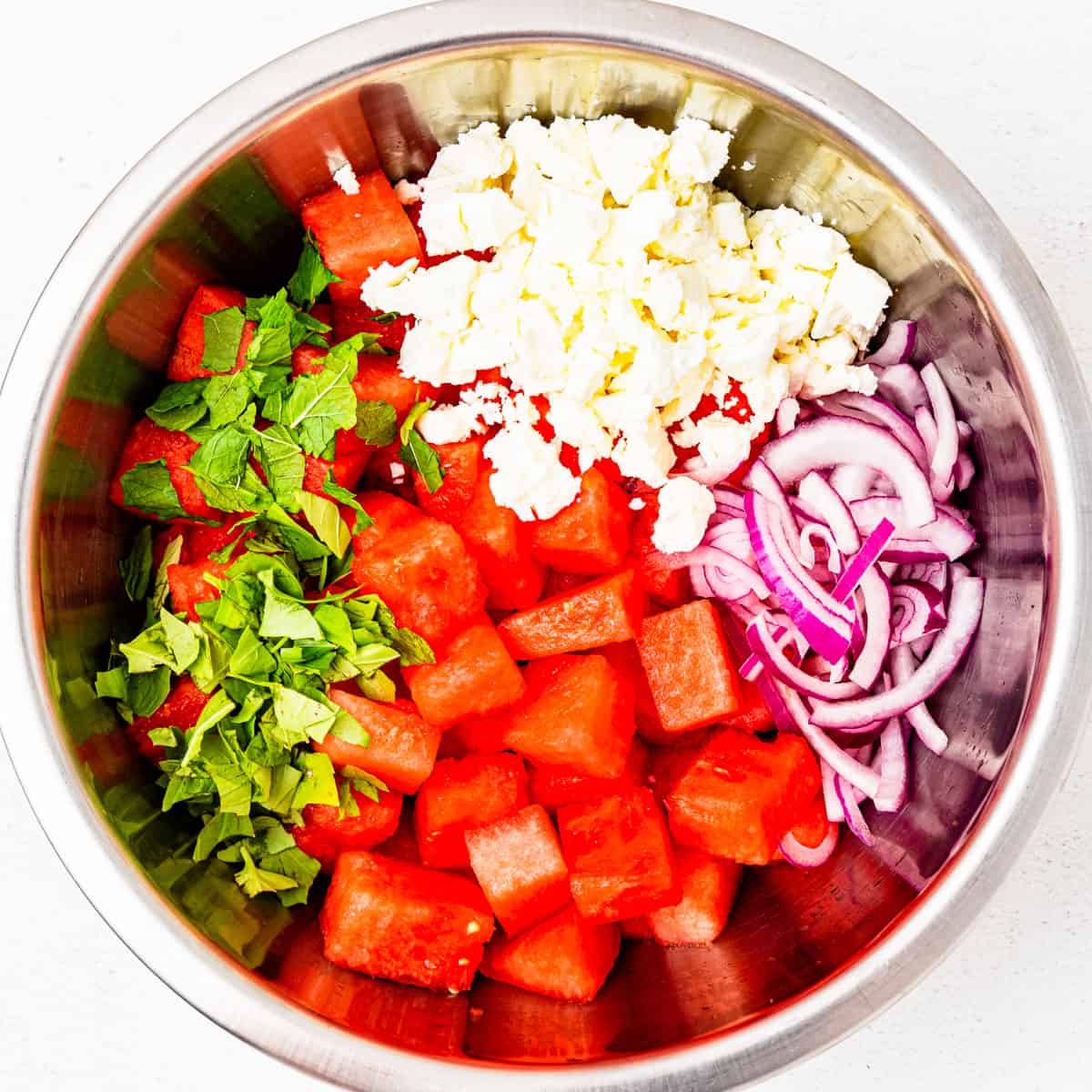 Cubed watermelon in a mixing bowl topped with feta cheese, red onion, and chopped herbs. 