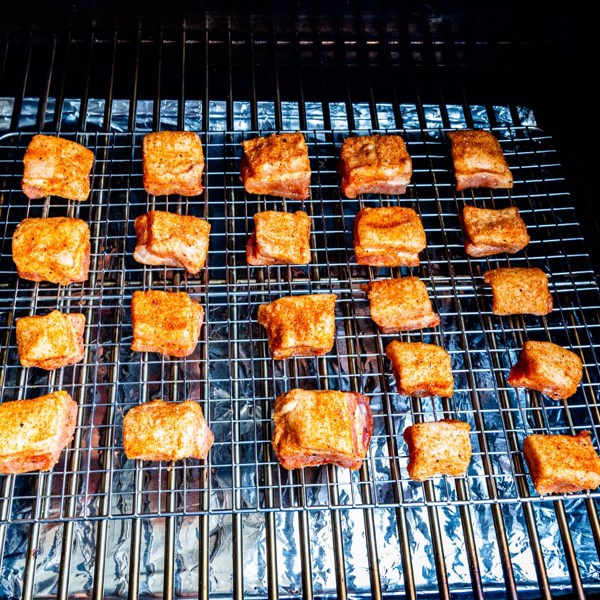 Seasoned cubes of pork belly cooking on a smoker.