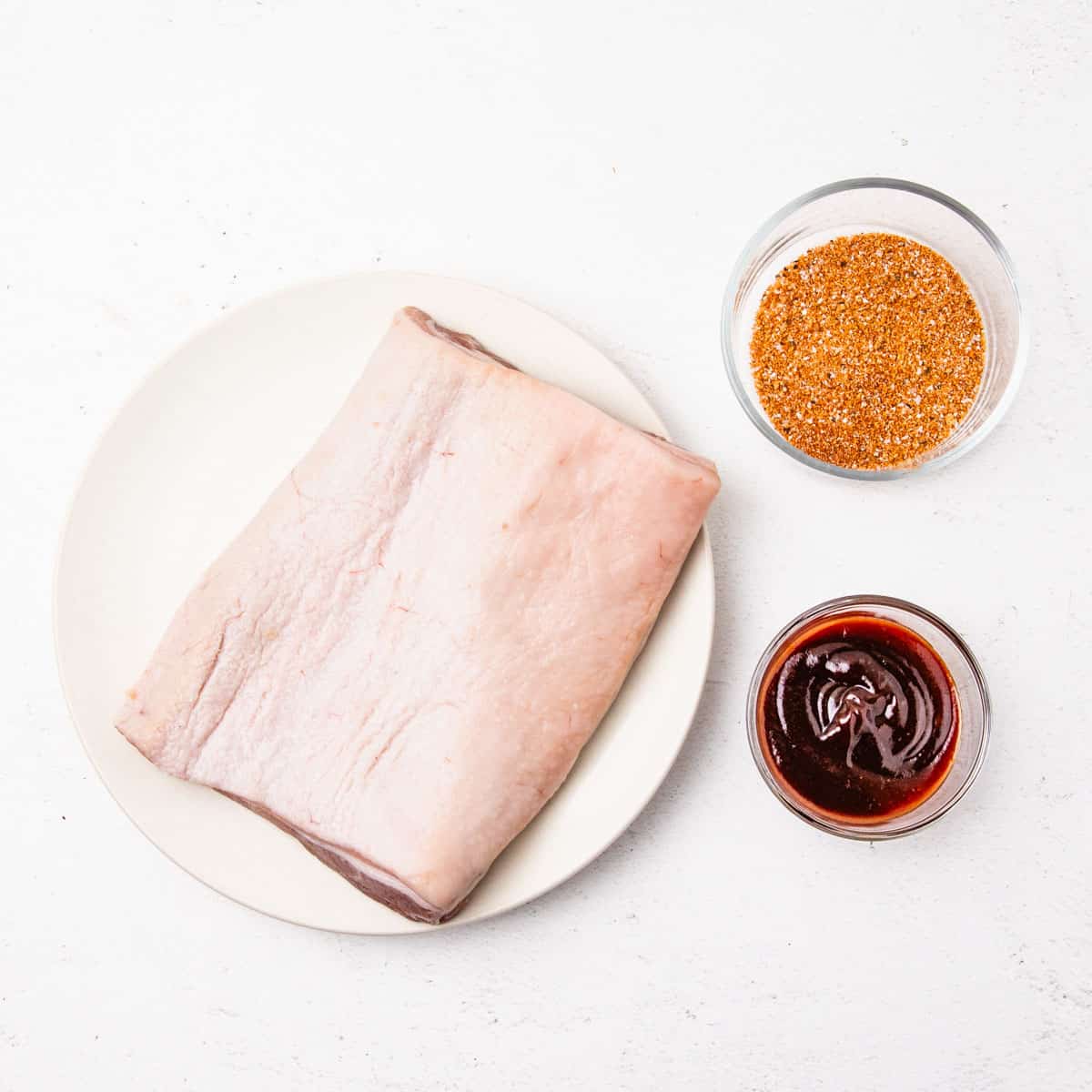 Small bowls of barbecue rub and barbecue sauce next to a slab of pork belly on a plate. 