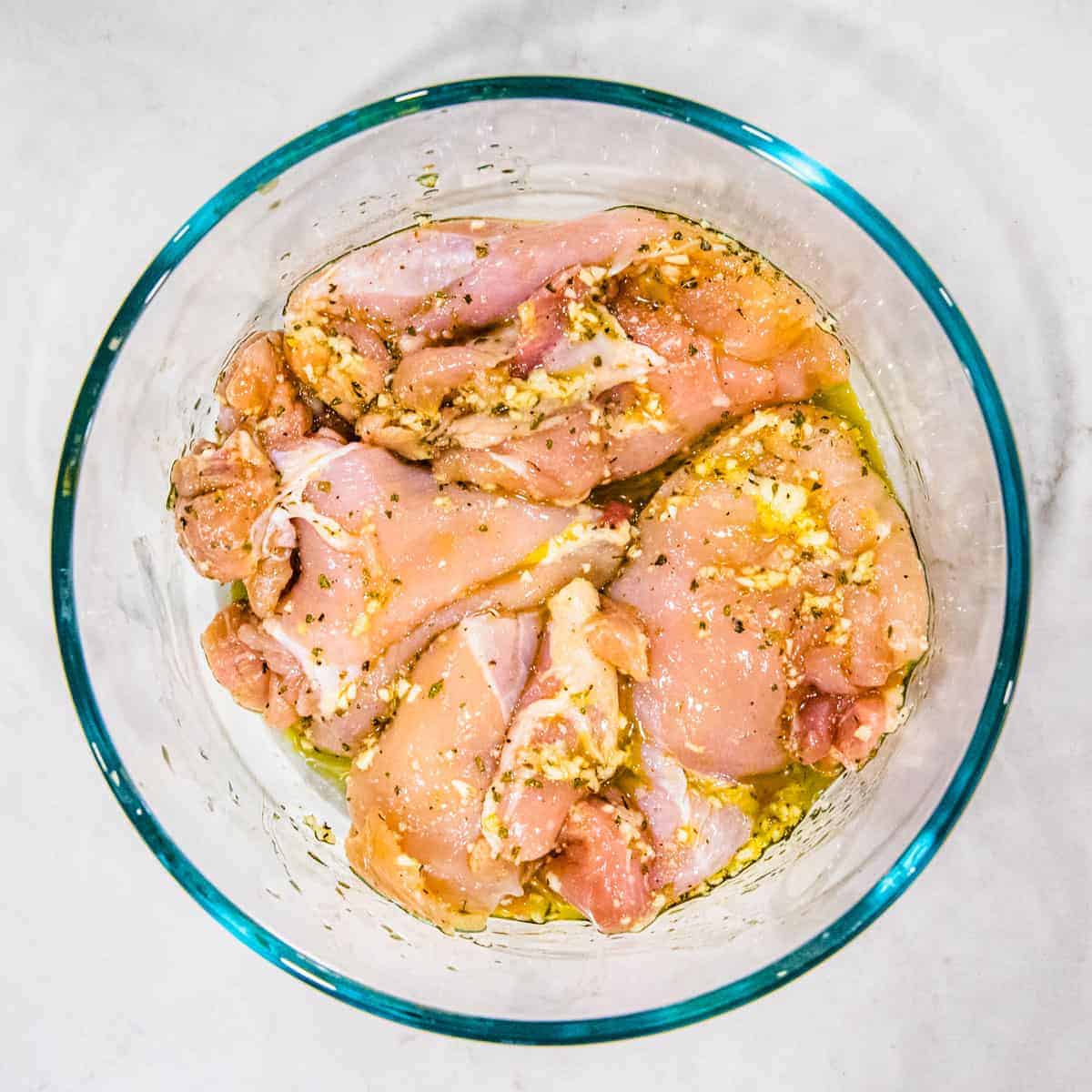 Chicken thighs in bowl with oil, lemon, and garlic seasoning.