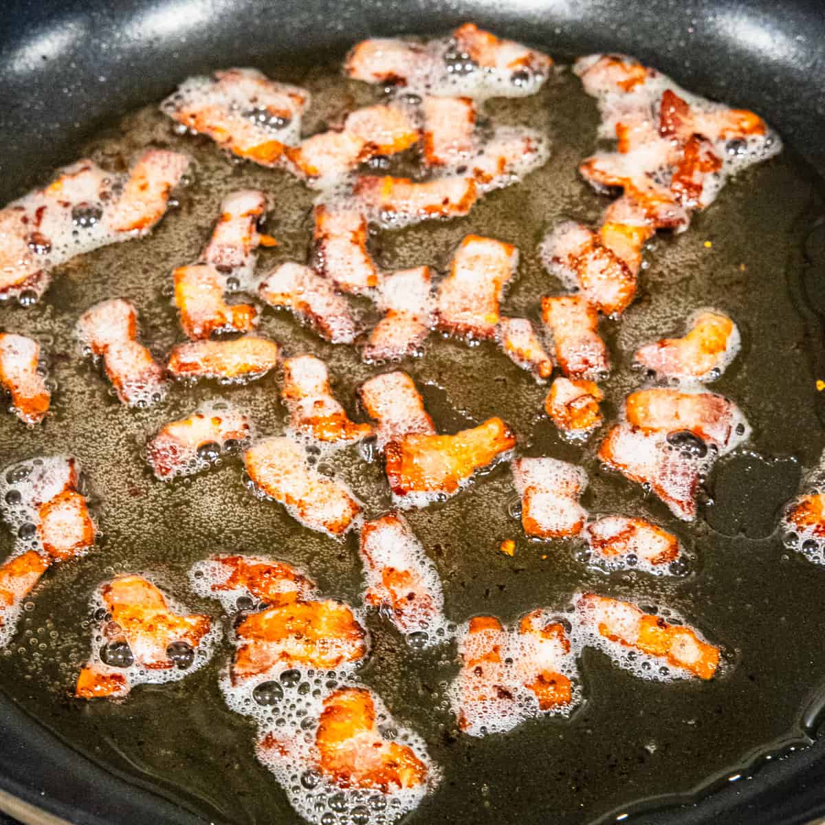 Chopped bacon strips being cooked in a large skillet.