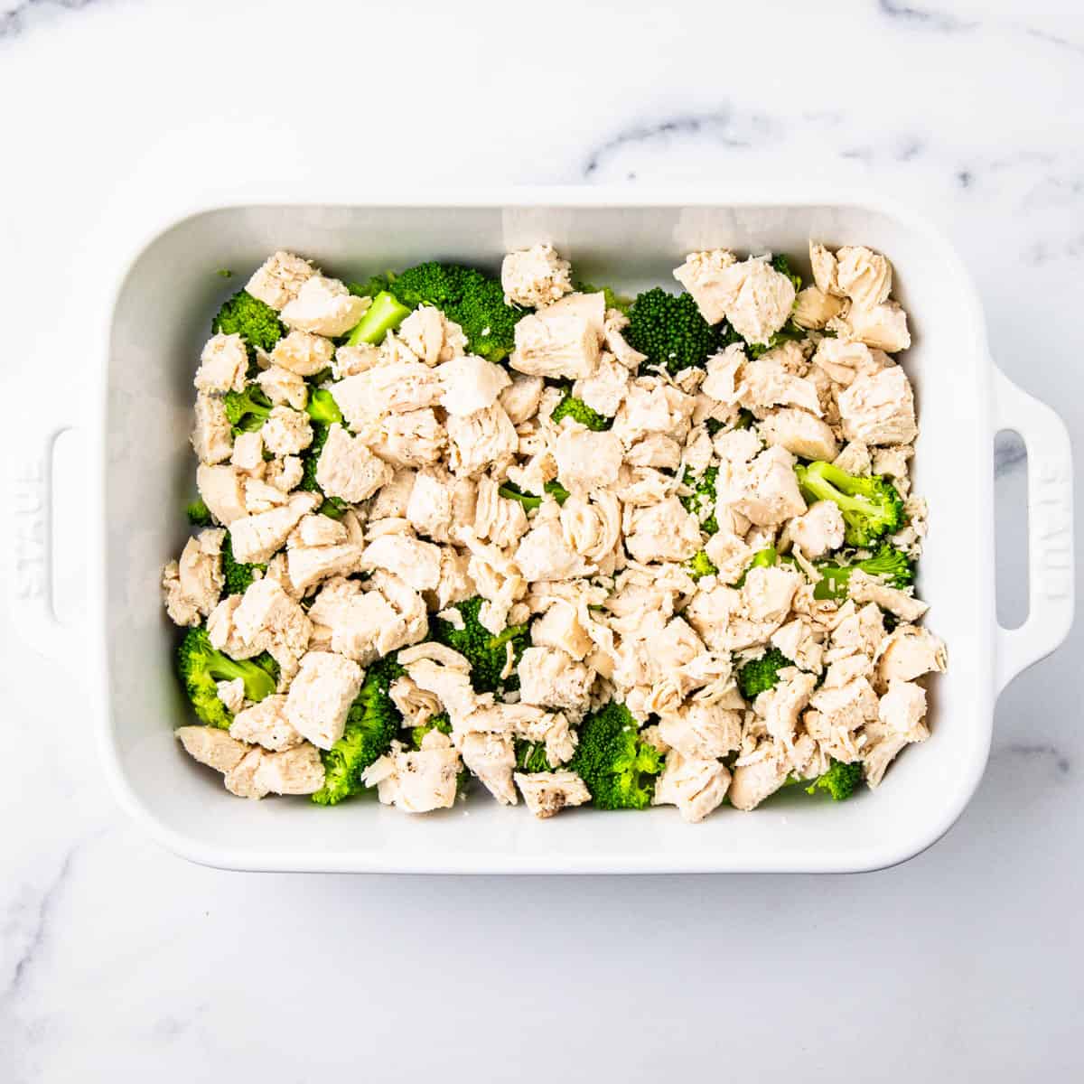 Cooked chicken spread over the top of broccoli in a baking dish.