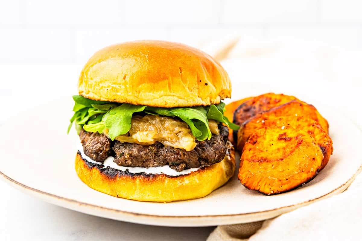 A lamb burger with caramelized onions and whipped feta sauce served on a plate with sweet potatoes.